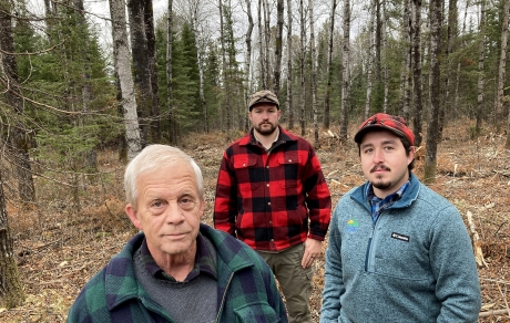 Three men stand in a clearing in the woods