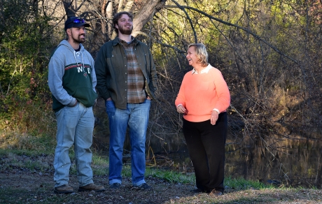 three people stand by a river with trees in the background