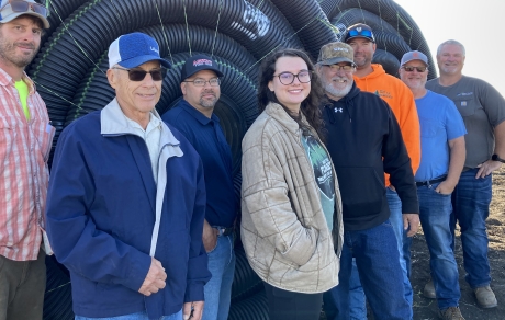 eight people stand in front of rolls of black plastic tile with a bare field in the background
